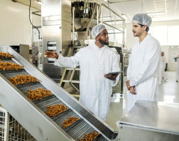 Diverse food factory quality control inspector with tablet talking to production line operator at facility.